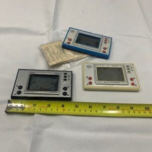 A group of three electronic game systems next to a ruler.